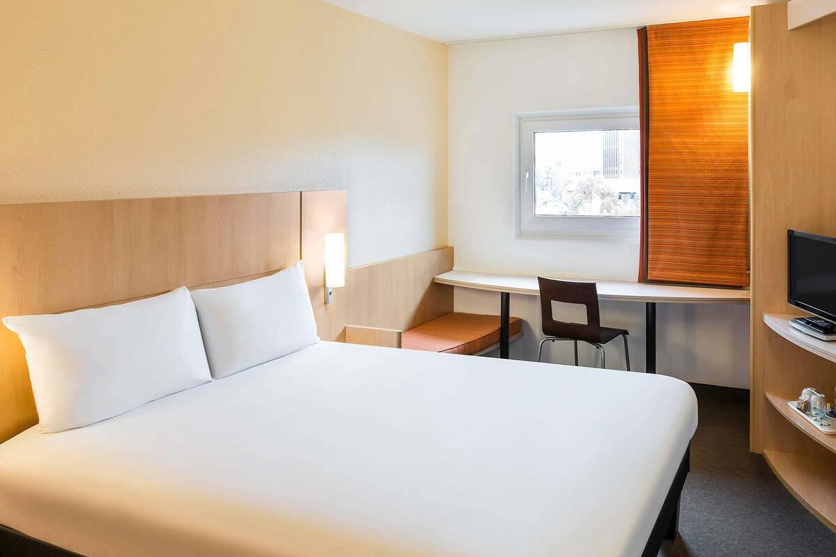 Ein Hotelzimmer mit einem großen Bett, einem Schreibtisch mit Stuhl, einem Fernseher und einem Fenster mit Jalousien.