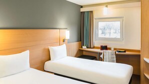 A hotel room with two beds, a desk, a chair, a window with a view of trees, and a lamp.