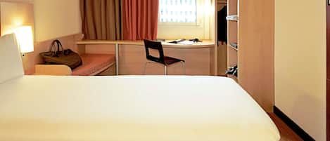 A hotel room with a large bed, a desk with a chair, a window with curtains, and a wardrobe.