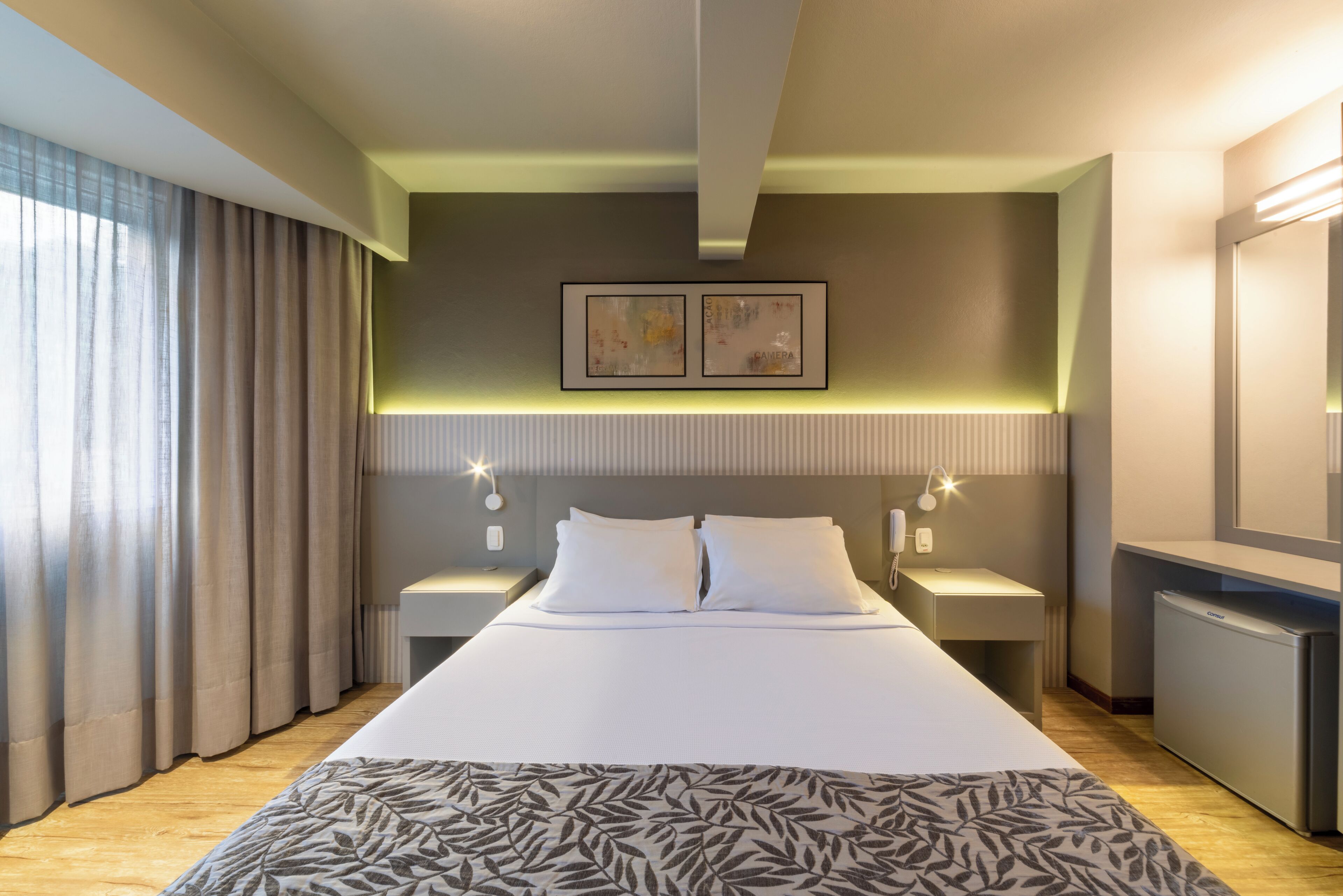 A hotel room with a large bed, two bedside tables with lamps, a small refrigerator, and a wall-mounted artwork.