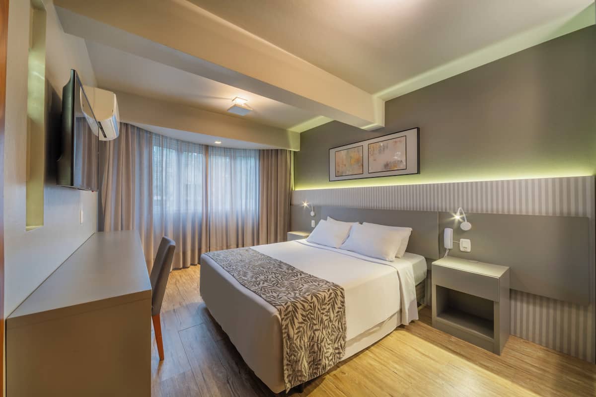 A modern hotel room with a large bed, bedside table, and a desk with a chair.