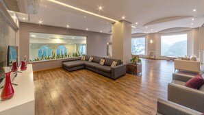 A spacious living area with a large sectional sofa, a glass wall with a cityscape view, and a wooden floor. - Hotel Laghetto Siena (Gramado)
