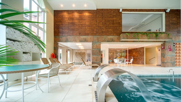 An indoor swimming pool with a waterfall feature, surrounded by white chairs and a table. - Hotel Laghetto Siena (Gramado)