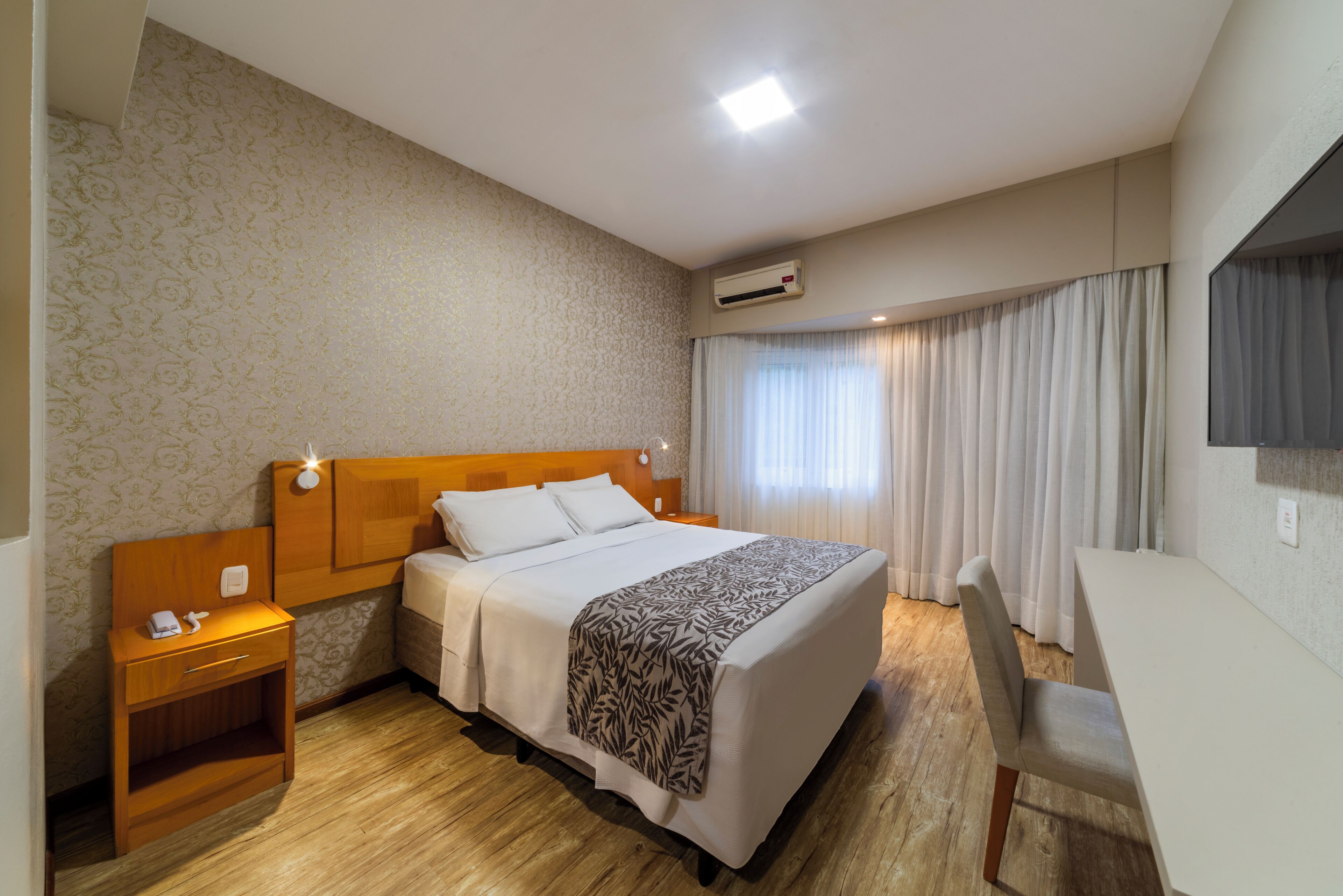 A hotel room with a bed, bedside table, chair, and an air conditioning unit.