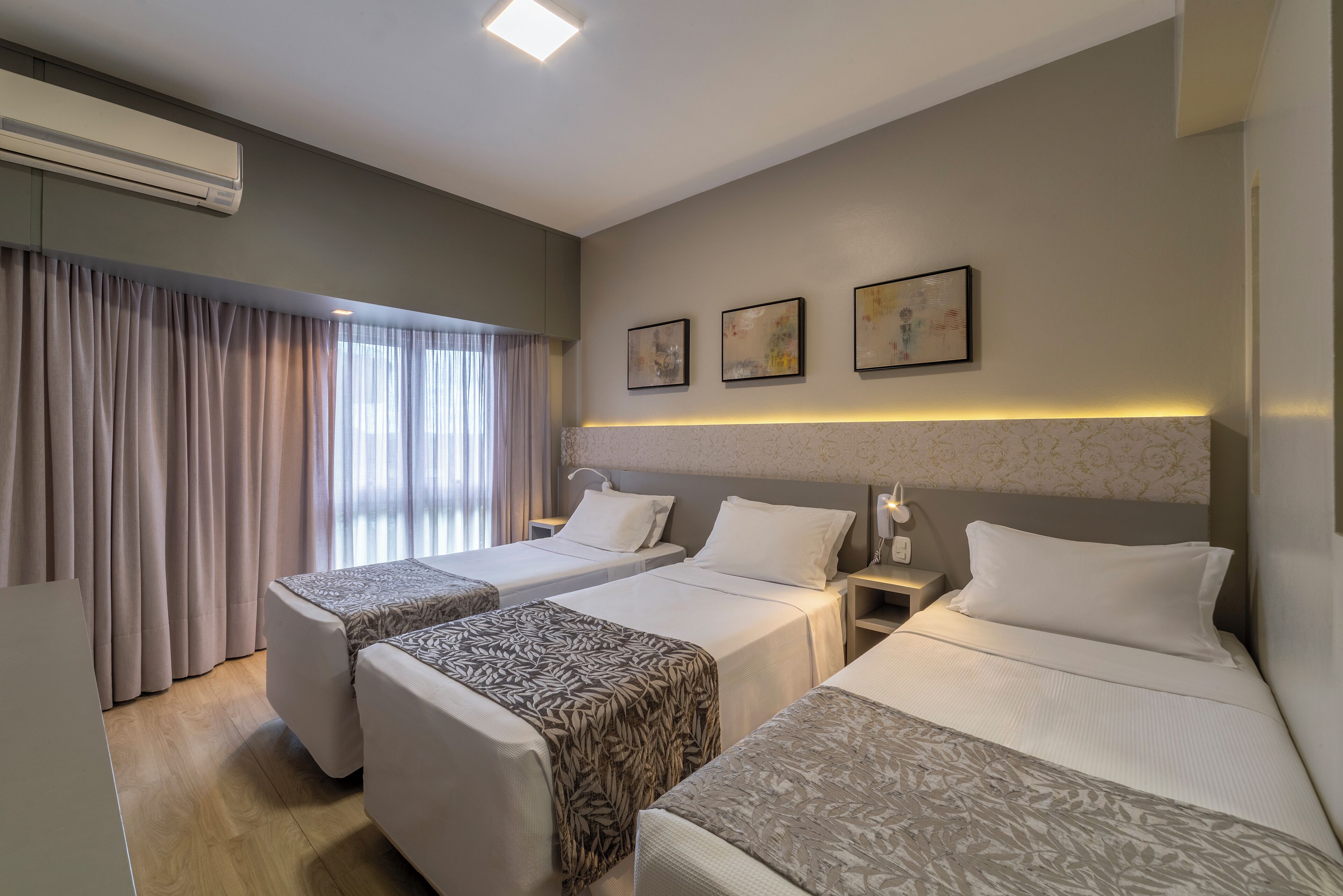 A hotel room with two single beds, a large window with curtains, an air conditioning unit, and three framed artworks on the wall.