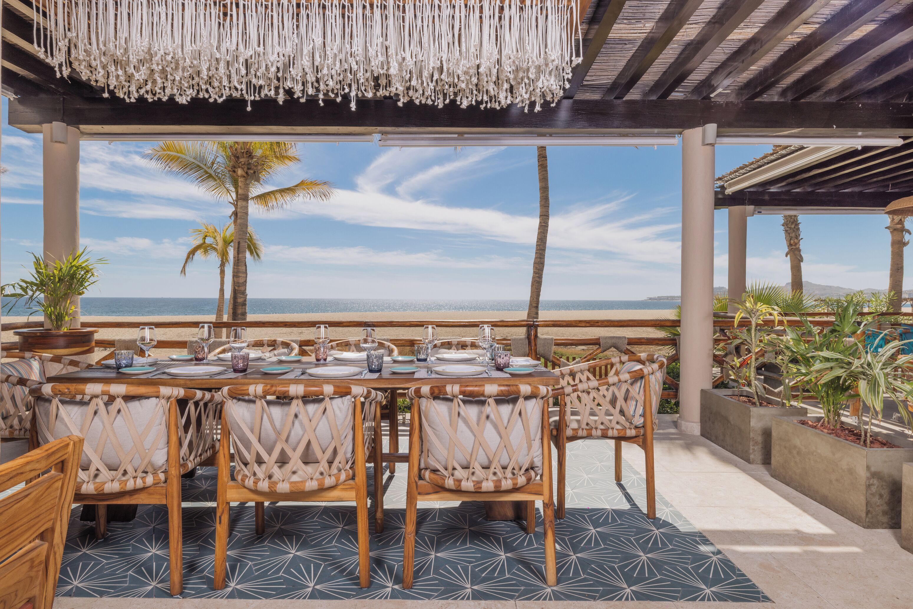 An outdoor dining area with a view of the ocean, featuring a round table with chairs, a hanging light fixture, and potted plants.