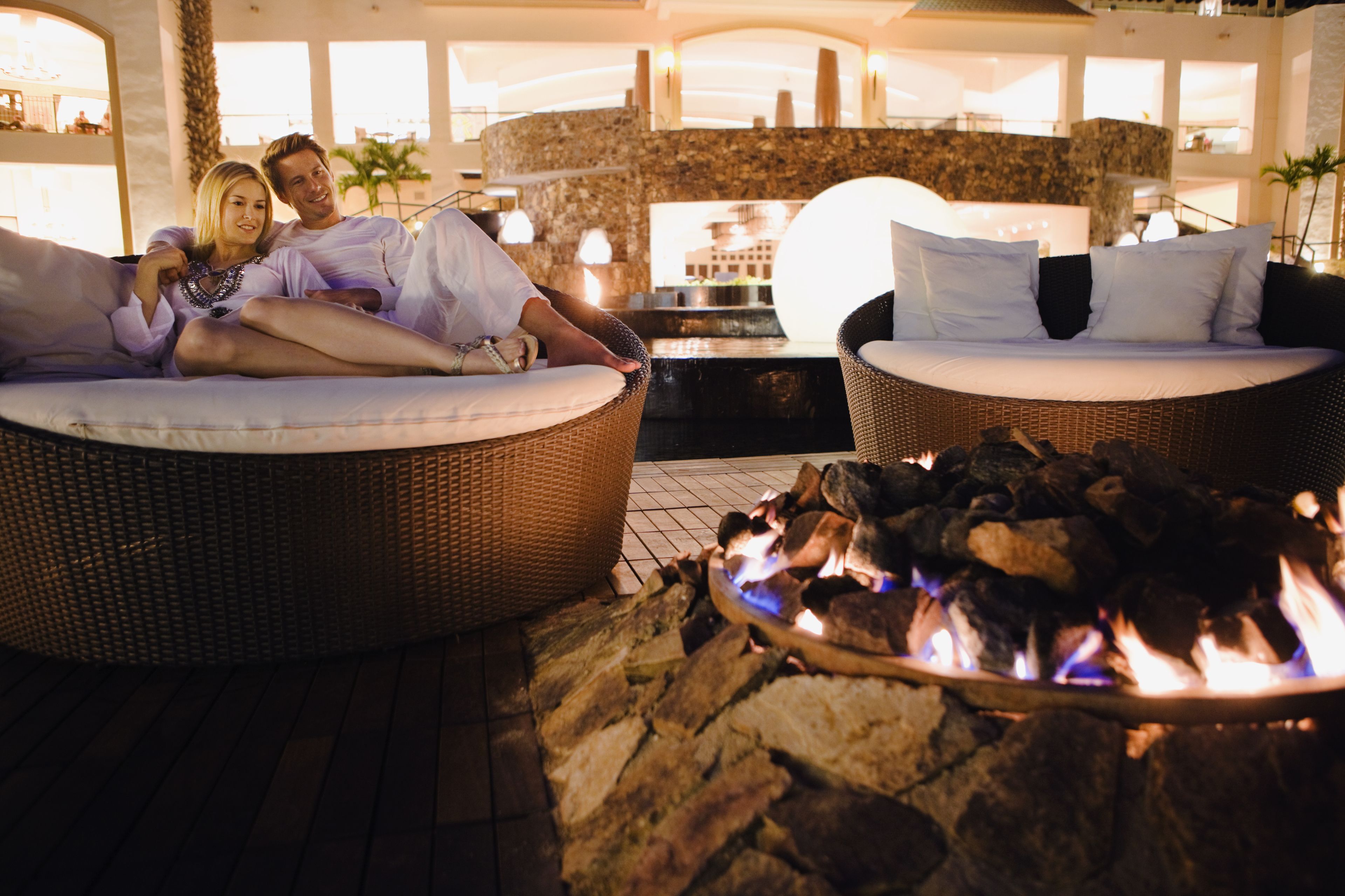 A couple relaxing on wicker sofas with white cushions, a stone fire pit in the foreground, and a view of a building through large windows.