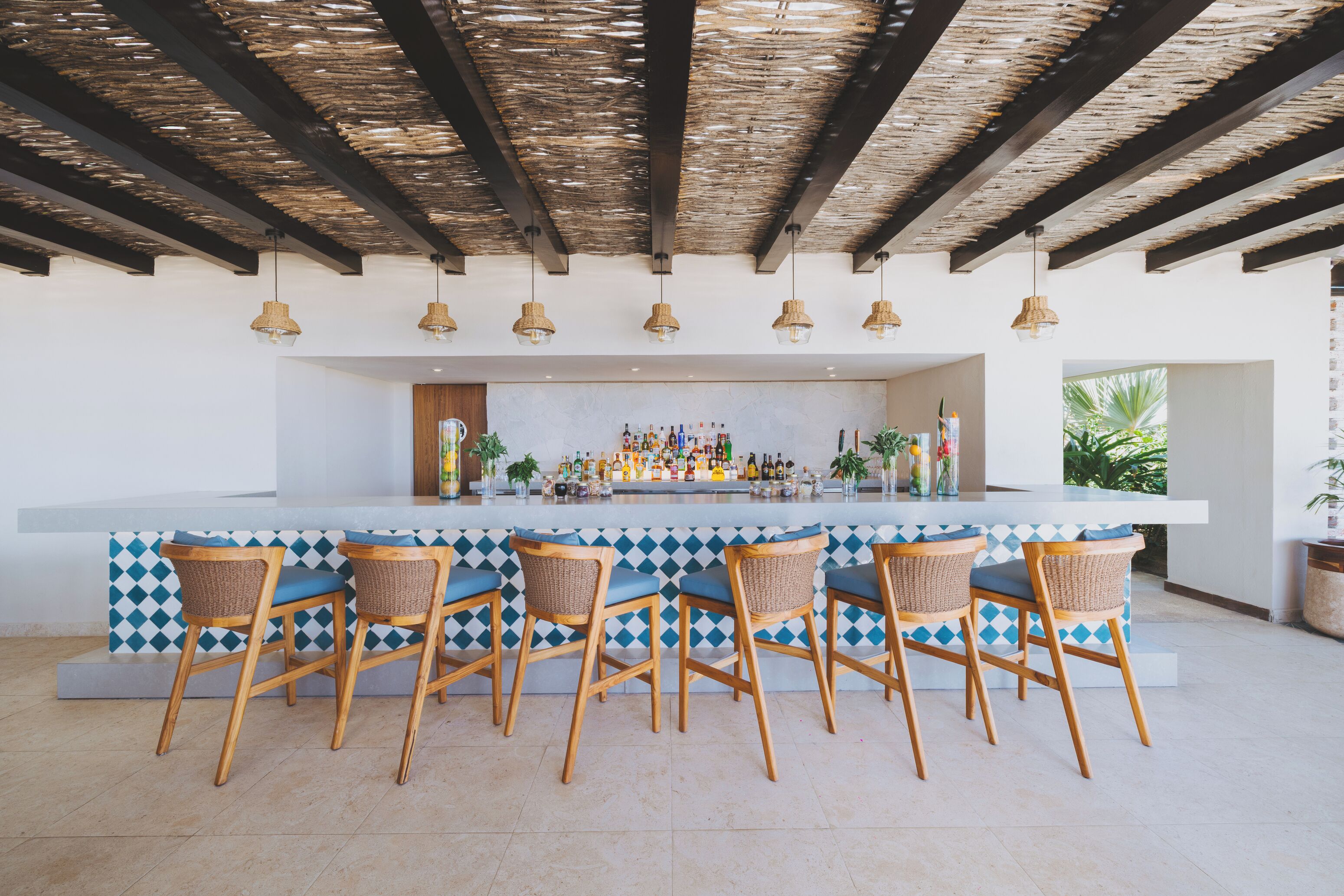 Un espace bar doté d’un comptoir blanc, de carreaux bleus et blancs, de chaises en osier et de luminaires suspendus.