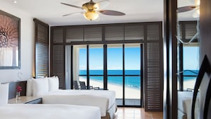 Quarto de hotel com duas camas, ventilador de teto e uma grande janela com vista para a praia.