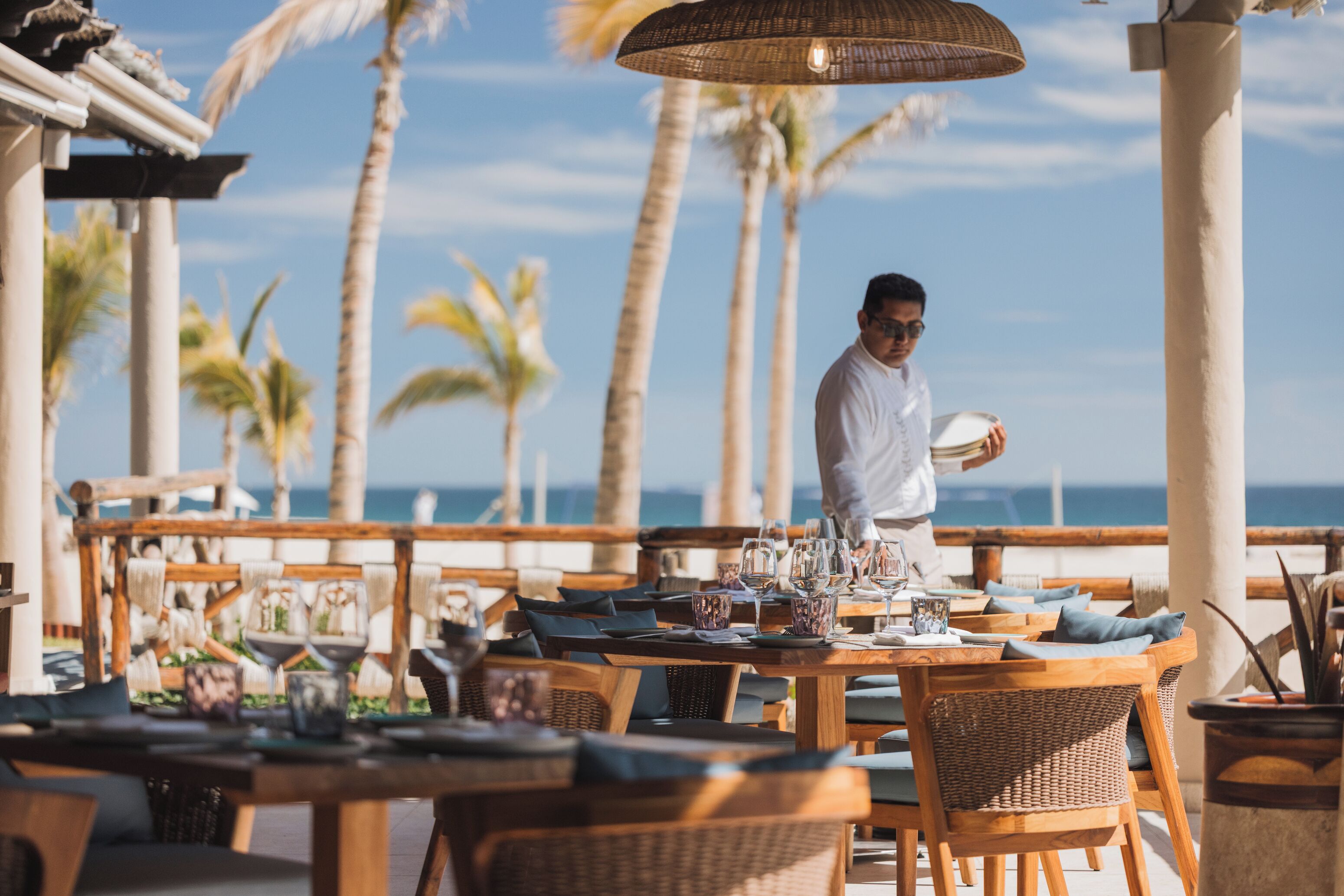A restaurant with outdoor seating, wooden tables, and chairs, overlooking a beach with palm trees.