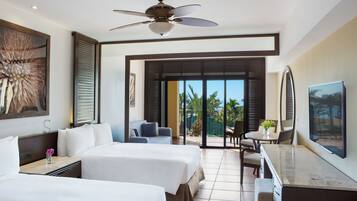 Habitación de hotel con dos camas, ventilador de techo, escritorio con silla, ventana grande y vista a palmeras.