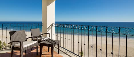 Un balcón con sillas de mimbre y una mesa, con vista a una playa y barandilla de metal.