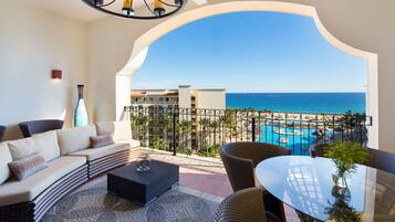 A balcony with a curved white arch, a sofa, wicker chairs, a glass table, and a view of the ocean and a pool.