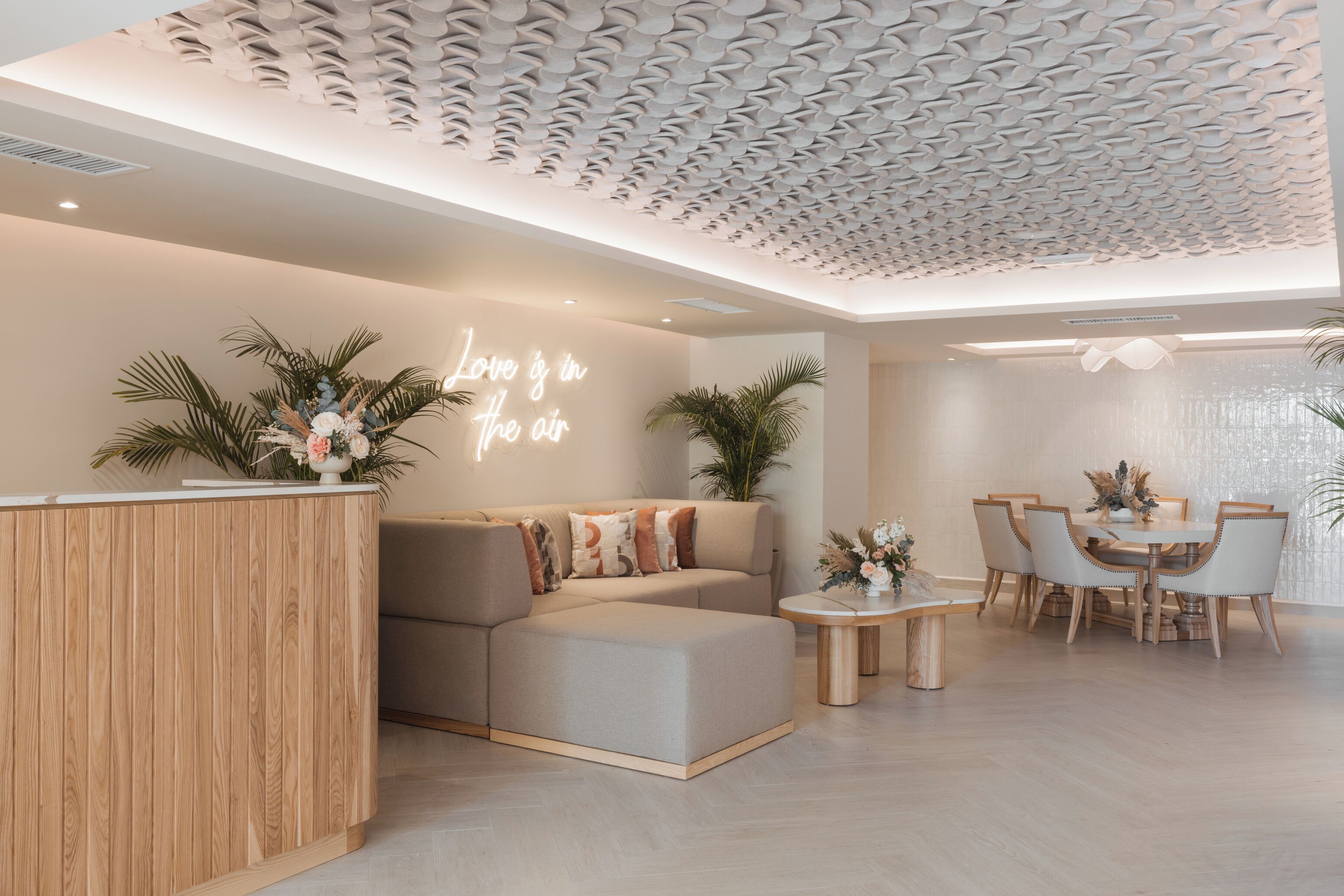 A modern living room with a sectional sofa, a wooden reception desk, a dining area with a round table and chairs, and a ceiling with a unique wavy pattern.