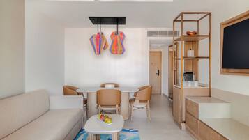 A modern living room with a sofa, a dining table with chairs, a small coffee table with fruit, a colorful pendant light, and a wooden bookshelf.