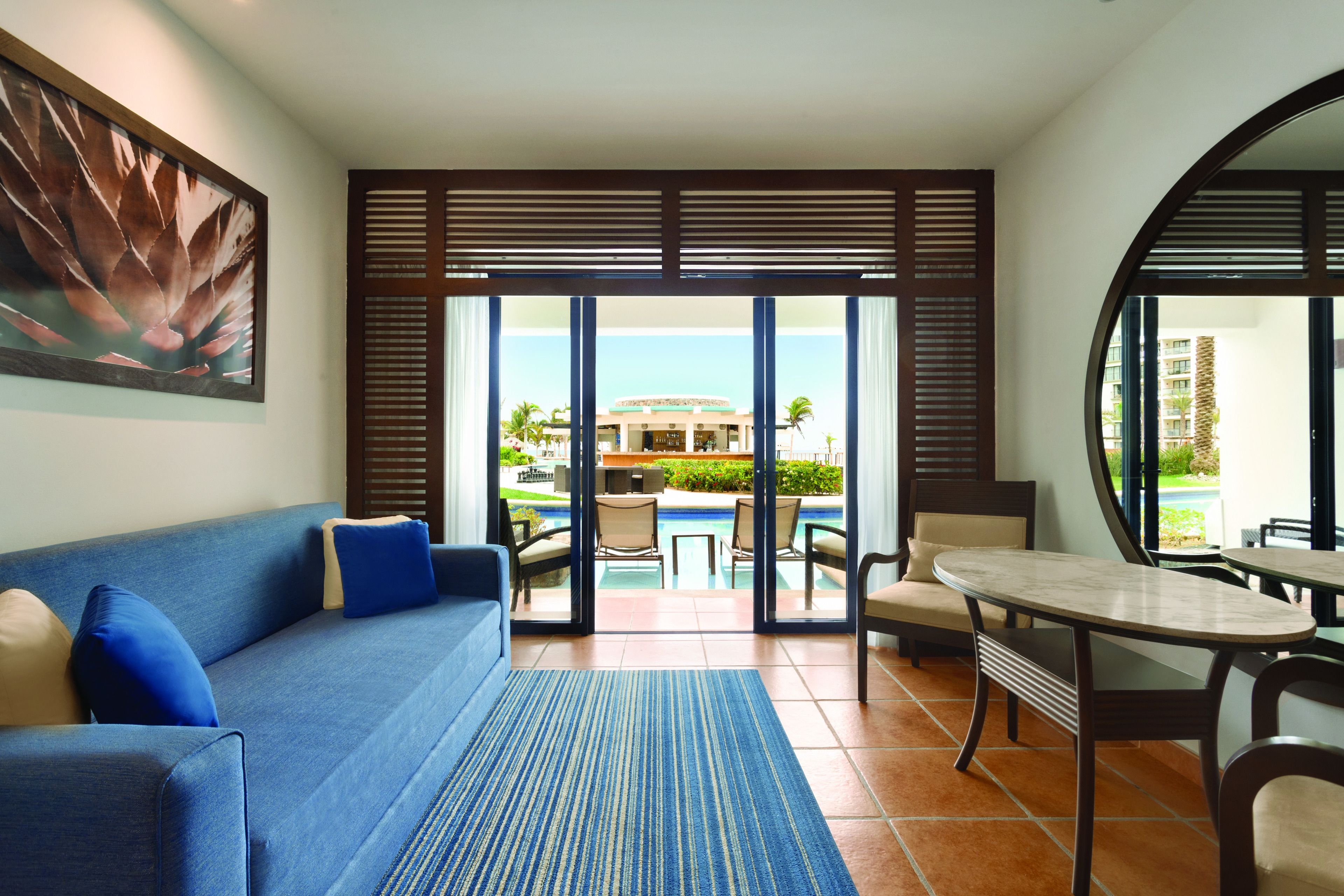 A modern hotel room with a blue sofa, a dining table, and a view of a pool and building.