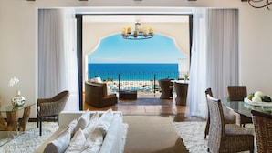 A modern living room with a glass table, wicker chairs, a sofa, and a view of the beach and ocean through a large window. - Hyatt Ziva Los Cabos- All Inclusive (San José del Cabo)