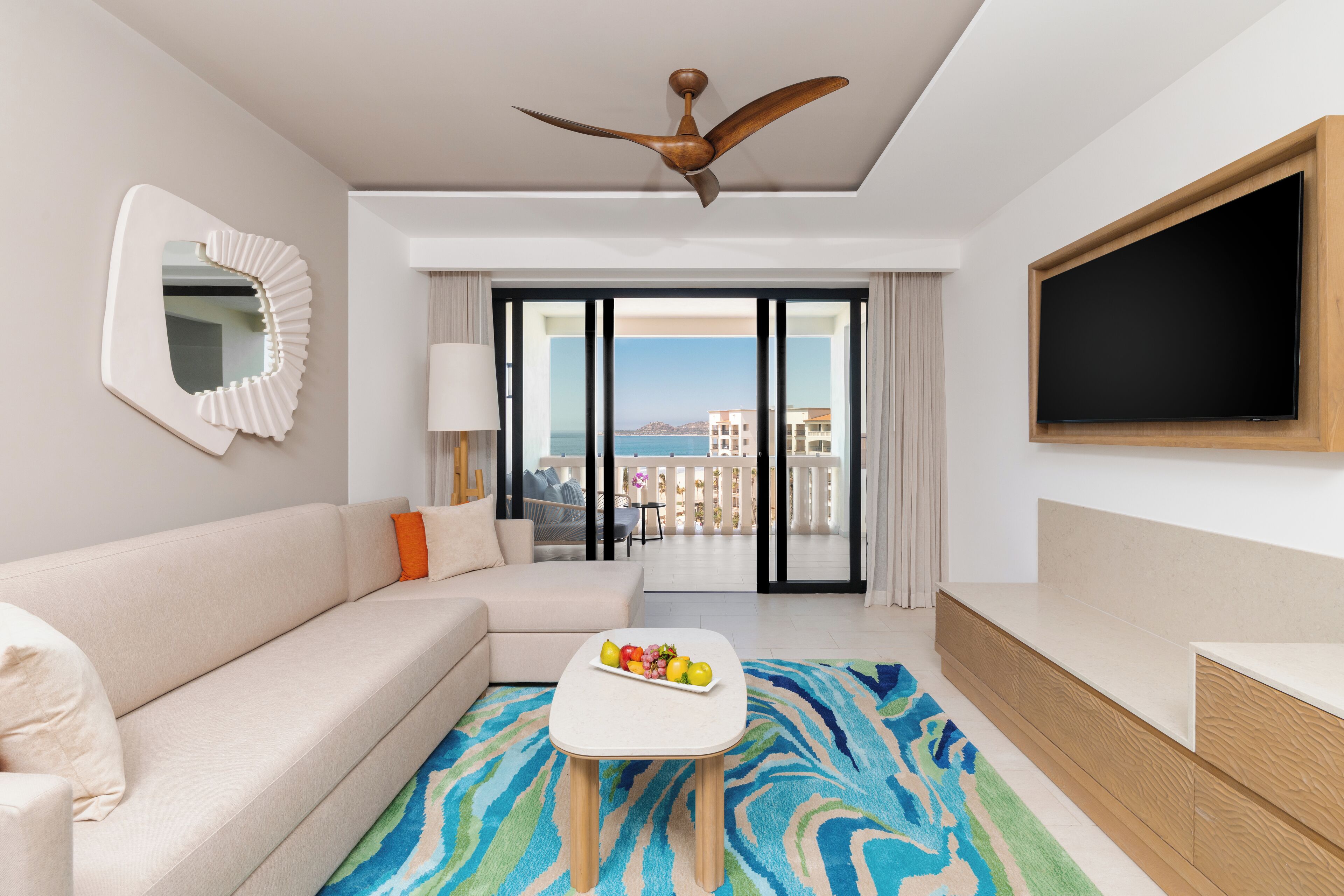 Une chambre d’hôtel moderne avec un canapé, une table basse sur laquelle sont disposés des fruits, un ventilateur de plafond, une télévision et un balcon avec vue.
