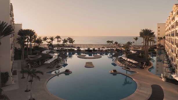Piscina de resort com design circular, cercada por palmeiras e áreas de estar, com vista para uma praia.