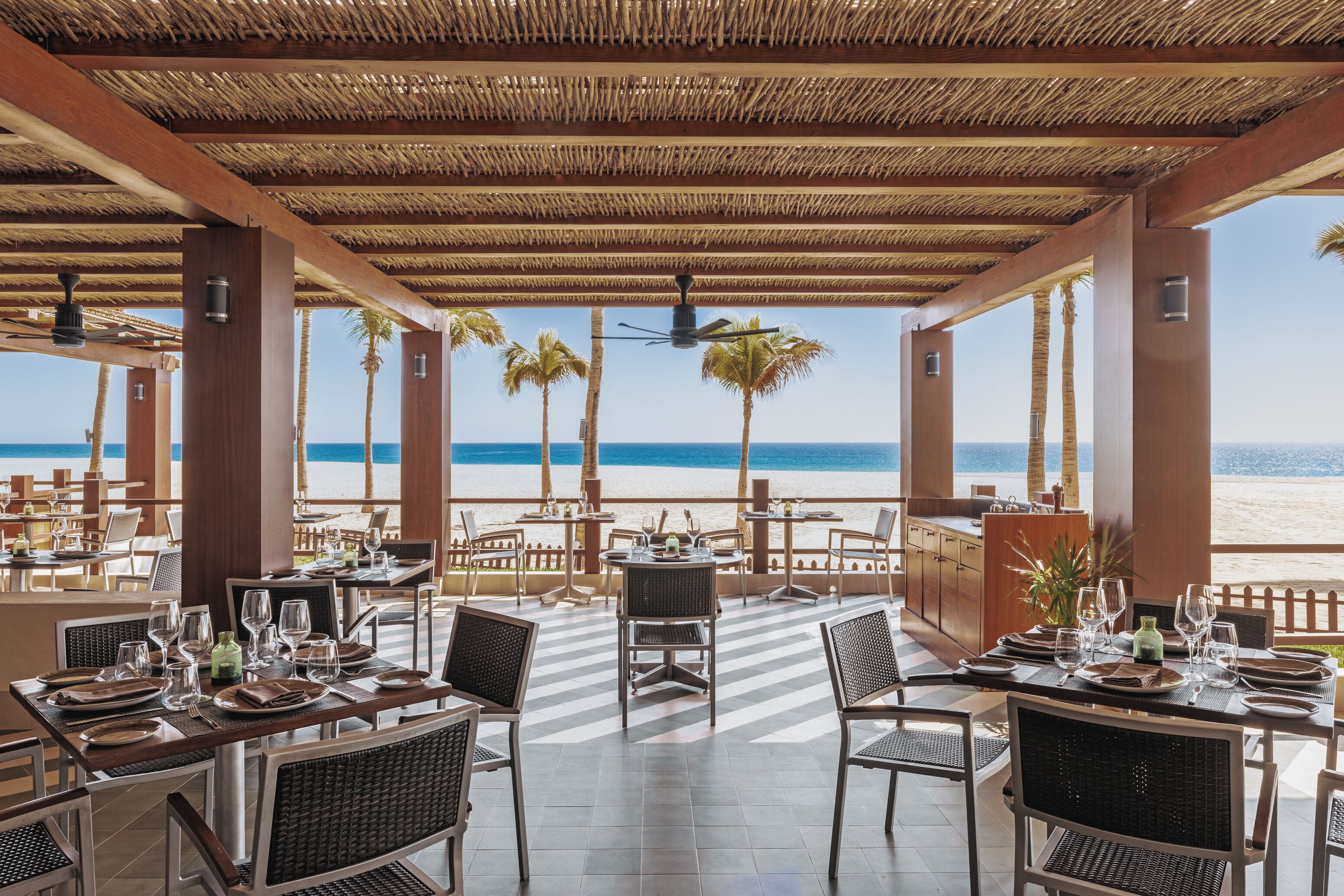 A beachside restaurant with tables set for dining, a thatched roof, and a view of the ocean.