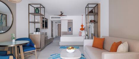 Una sala de estar moderna con sofá, mesa de centro y sillas azules. Se observa una zona de cocina con refrigerador, microondas y estanterías. Se distingue un ventilador de techo, y la habitación cuenta con un espejo grande y un florero decorativo.