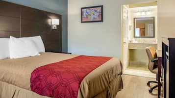 Habitación de hotel con cama, manta roja, cabecero, lámpara de pared, mapa enmarcado, baño con espejo y escritorio con silla.