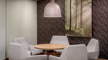 Une salle de conférence avec une table ronde et quatre chaises, un luminaire suspendu et un grand tableau représentant une forêt, accroché au mur.