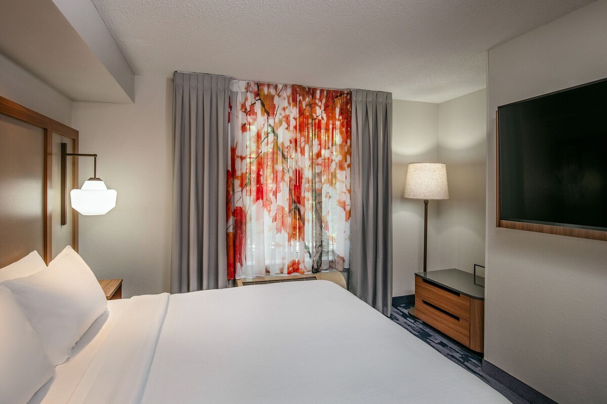 Ett hotellrum med en säng, ett skrivbord, en lampa och ett stort fönster med gardiner.
