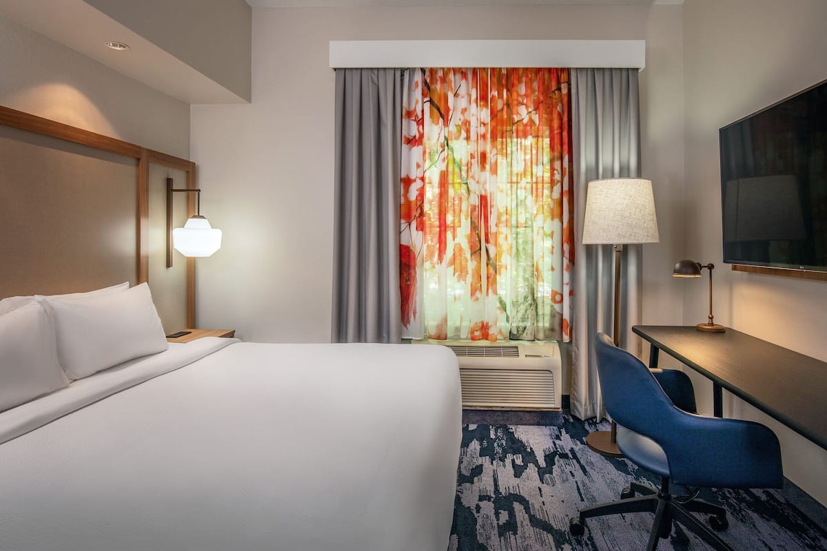 Habitación de hotel con cama, escritorio, silla, televisor, lámpara y ventana con cortinas.
