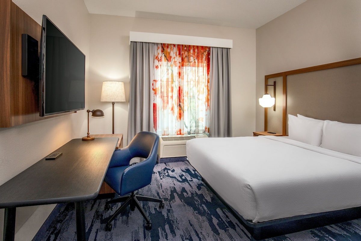 Habitación de hotel con cama, escritorio, silla, televisor, lámpara y ventana con cortinas.