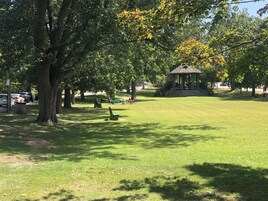 A park with a gazebo, green grass, trees, and people sitting on benches.