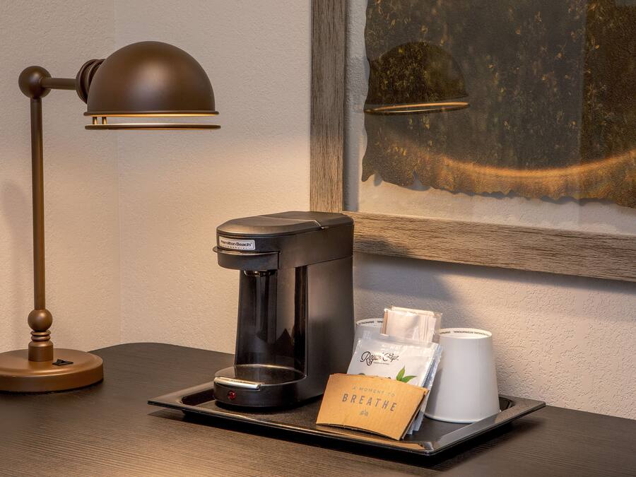 Una cafetera, una bandeja con una taza de café y una bolsa, y una lámpara sobre una mesa de madera.