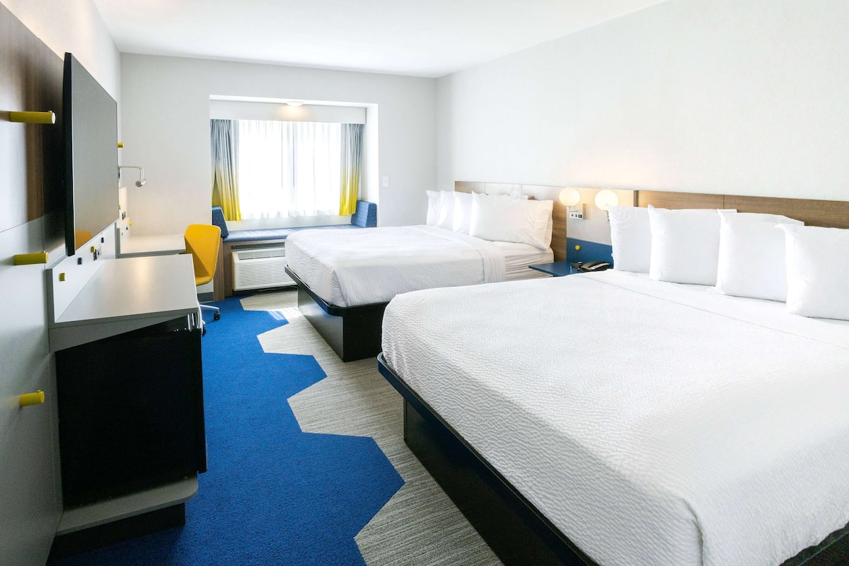 Quarto de hotel com duas camas, uma escrivaninha, uma cadeira, uma televisão e uma janela com cortinas.