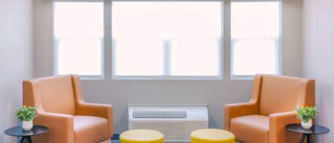 Une pièce avec deux fauteuils orange, un climatiseur blanc et deux poufs jaunes, le tout posé sur un tapis à motifs bleus et gris.