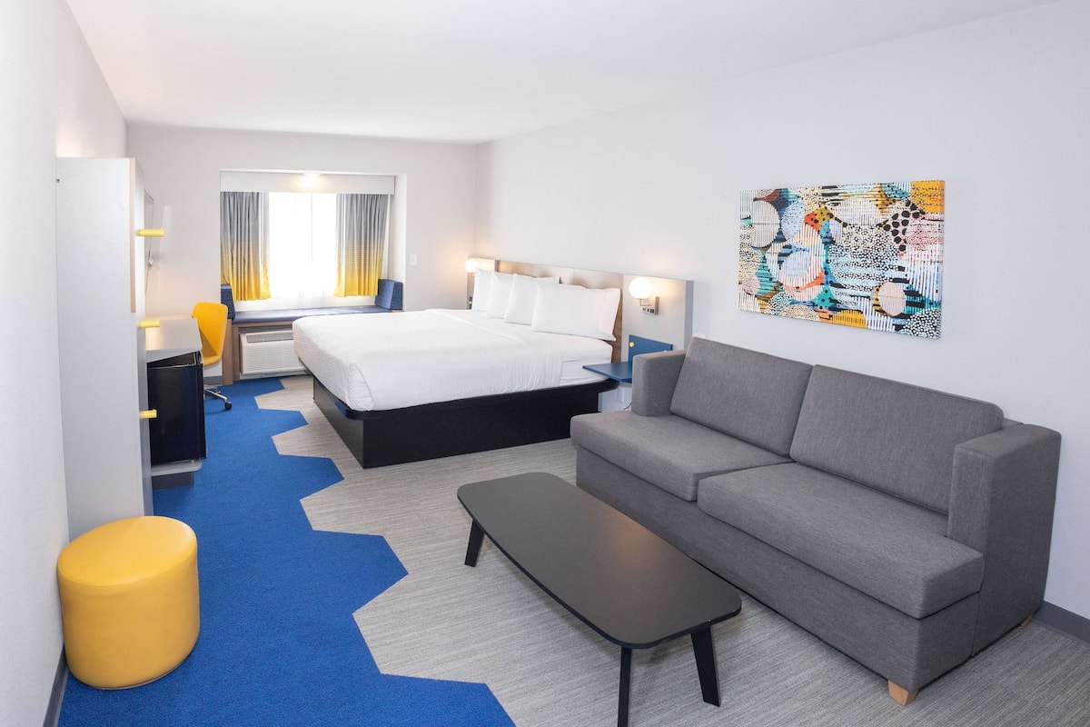 Quarto de hotel moderno com cama, sofá cinzento, pequena mesa e um banquinho amarelo.