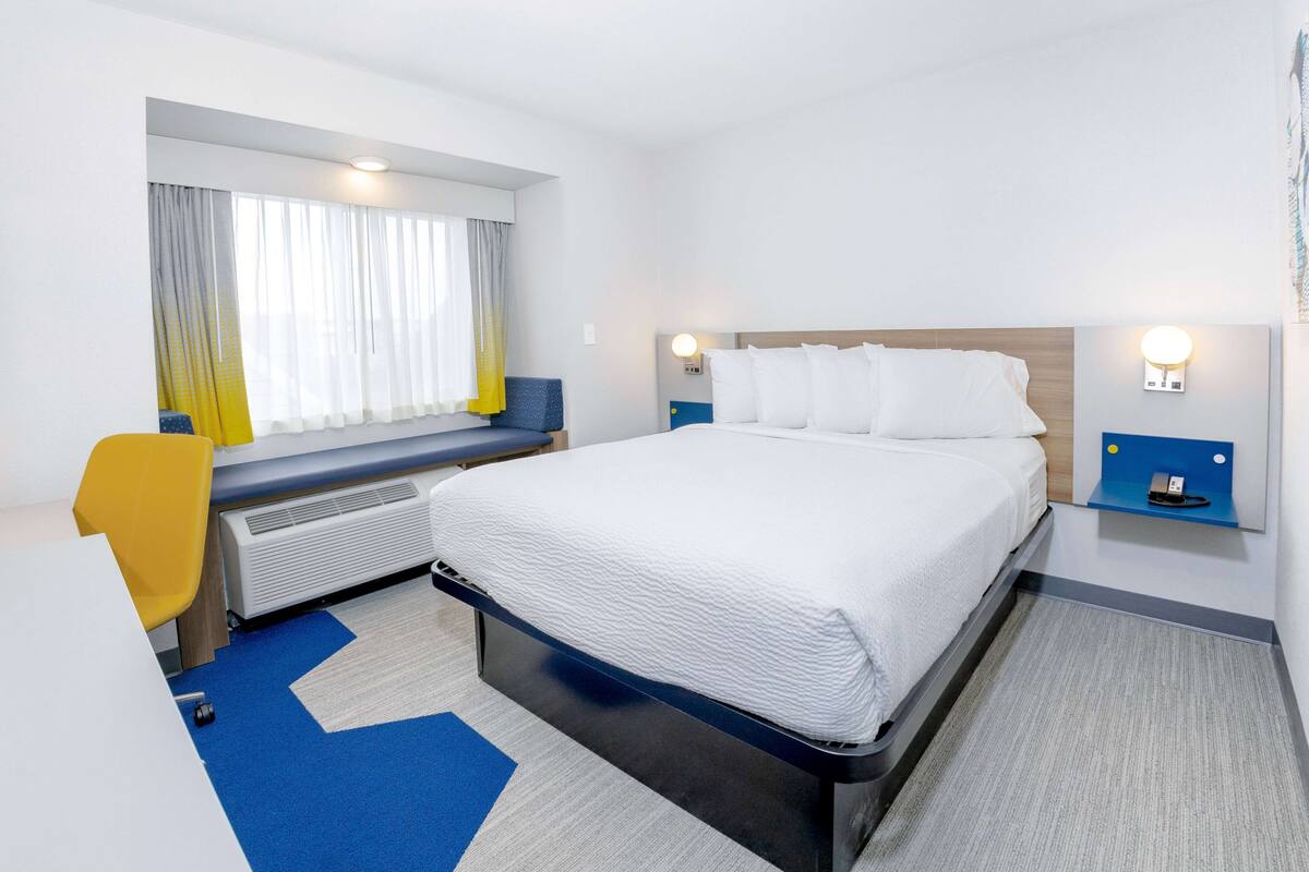 Quarto de hotel com cama, escrivaninha, cadeira, janela com cortinas e mesa de cabeceira com abajur.