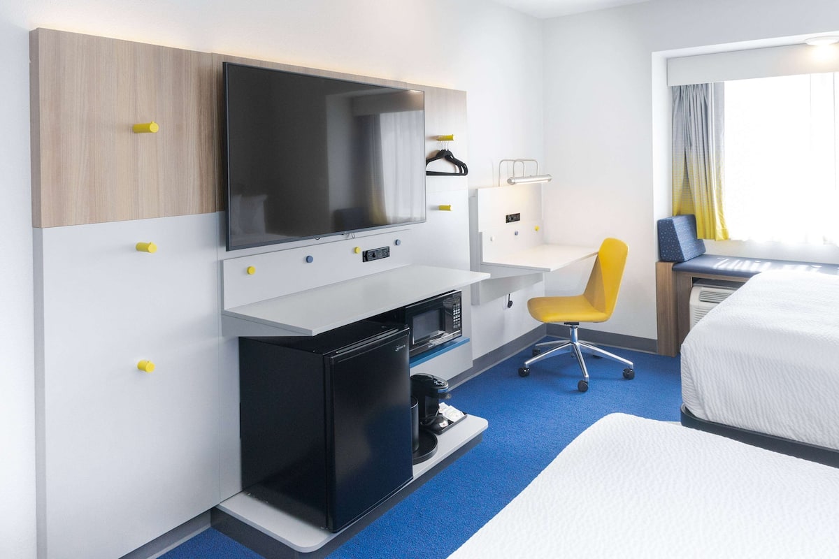 Quarto de hotel com cama, escrivaninha com computador, televisão, mini-geladeira e uma cadeira.