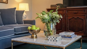 Een zithoek met een bank, een bijzettafeltje met een schaal appels, een vaas met bloemen en een stapel boeken.