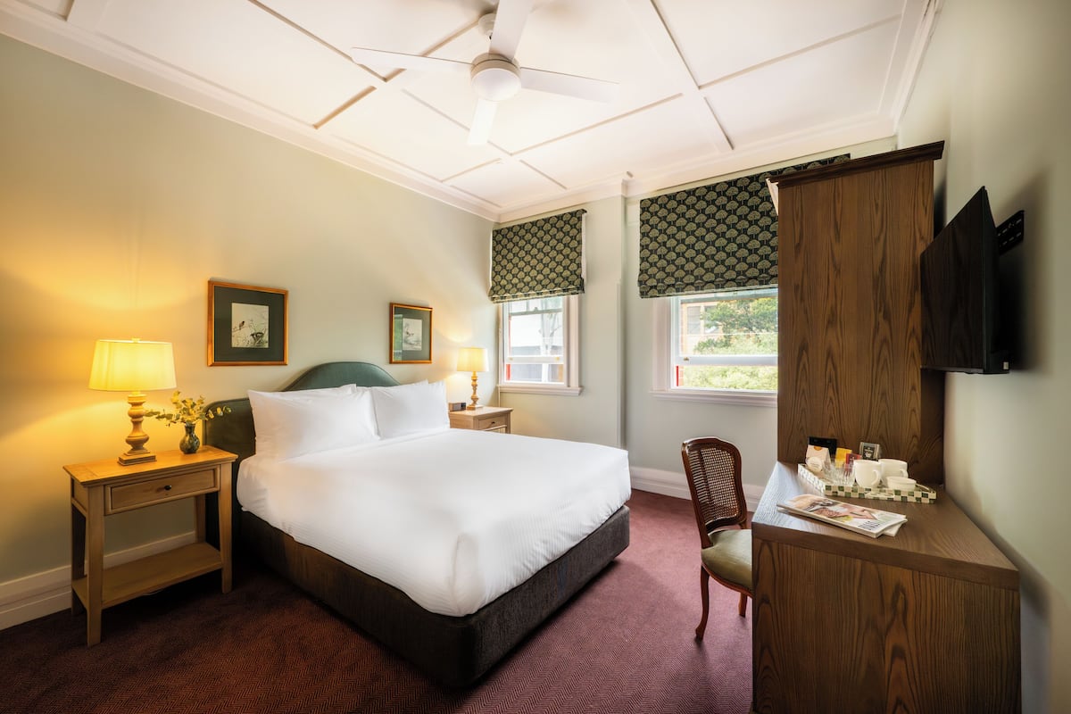 A hotel room with a large bed, a desk with a chair, a lamp, and framed pictures on the wall.