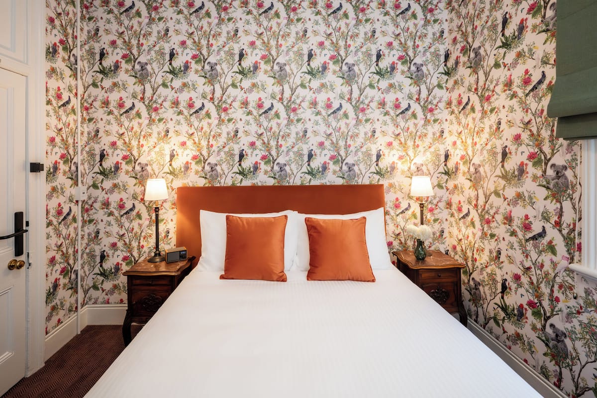 A bedroom with a floral wallpaper, a large bed with two pillows, two bedside tables with lamps, and a door.