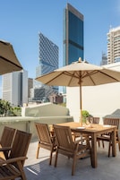 An outdoor dining area with wooden chairs and a table, surrounded by city buildings.