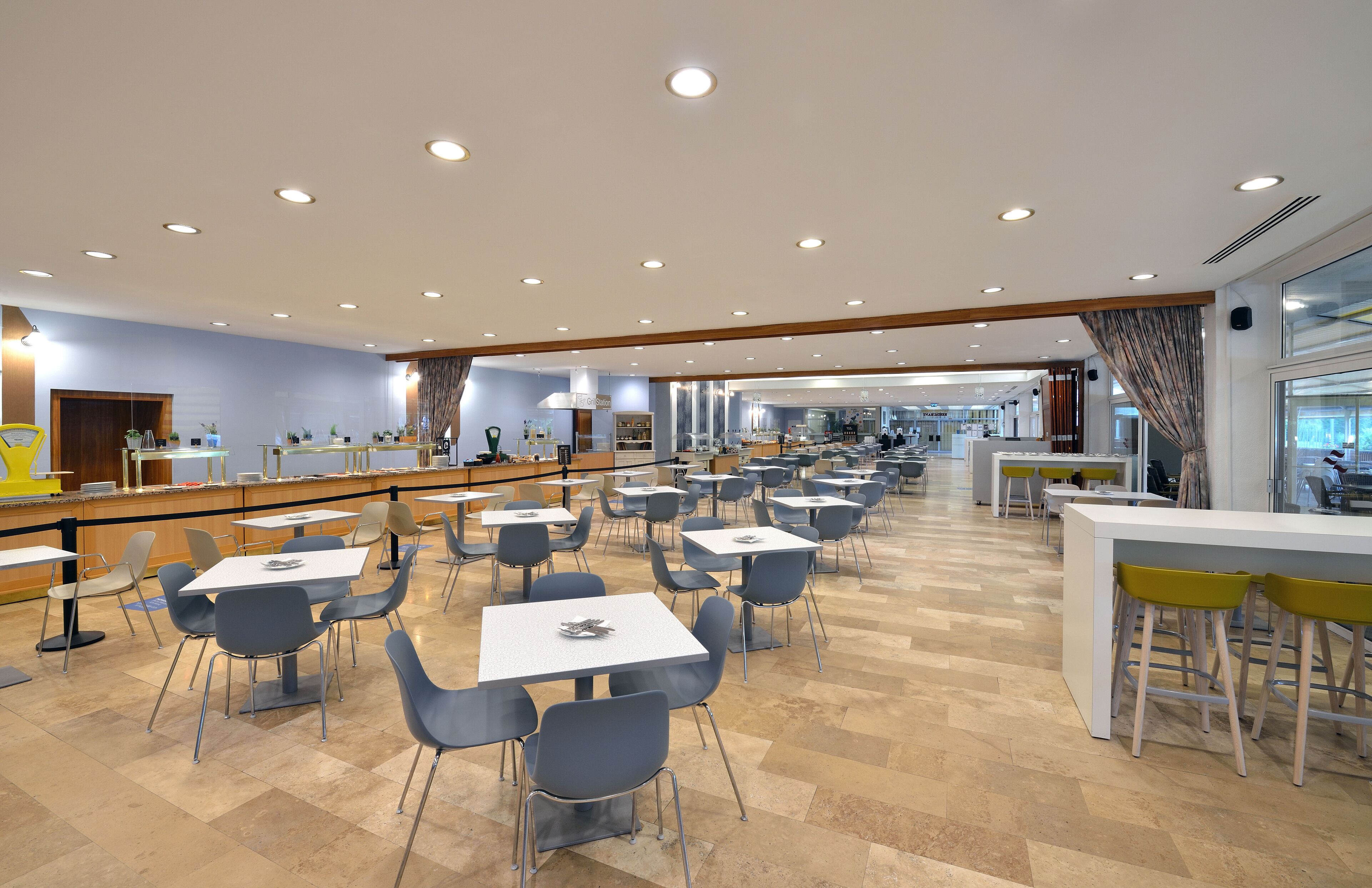 Eine großzügige Cafeteria mit mehreren Tischen und Stühlen, eine Theke mit einem gelben Stuhl und ein großes Fenster auf der rechten Seite.