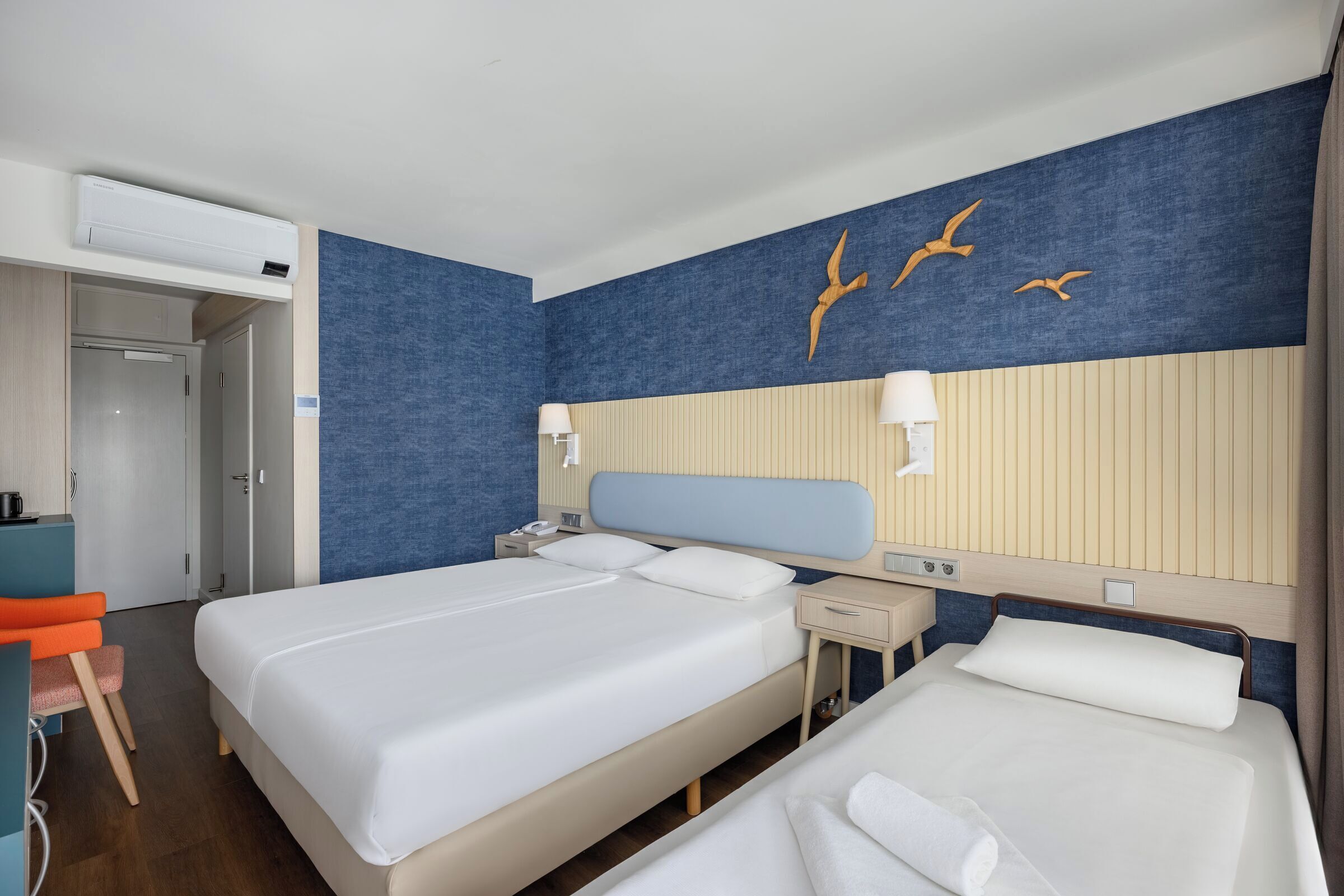 Een hotelkamer met twee bedden, een bureau en een stoel. Daarboven hangt een airconditioner en aan de muur is een vogelmuurdecoratie te zien.