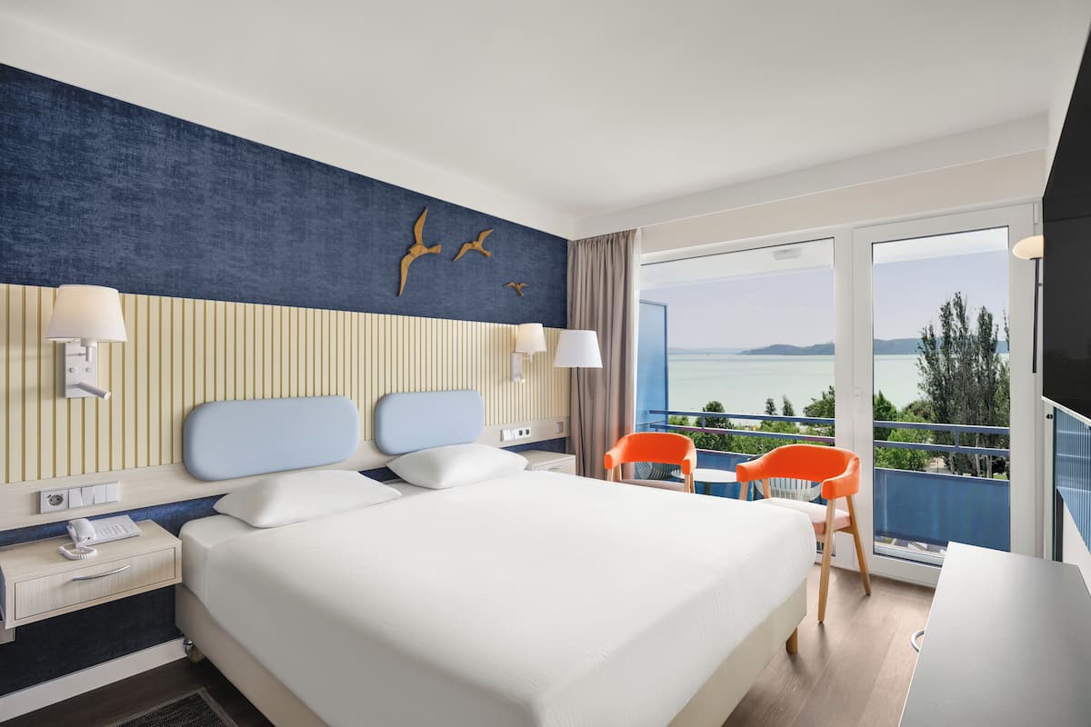A hotel room with a large bed, two bedside tables, a desk, and a balcony with a view of the sea.