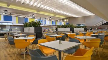 Un espace de restauration moderne avec plusieurs tables et chaises, une cuisine en arrière-plan et un plafond doté de luminaires linéaires.