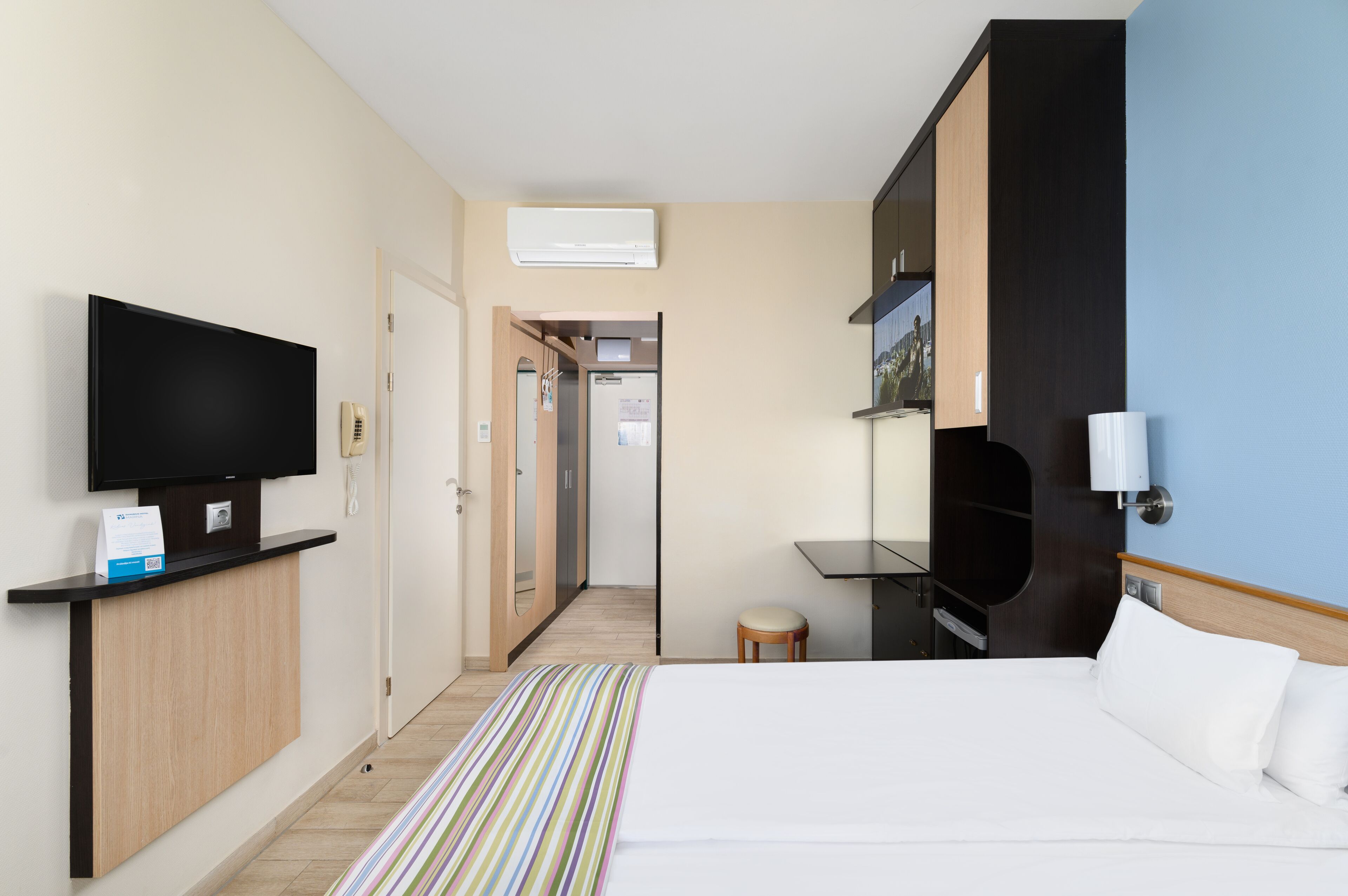 A hotel room with a bed, a TV, a desk, and a small stool.
