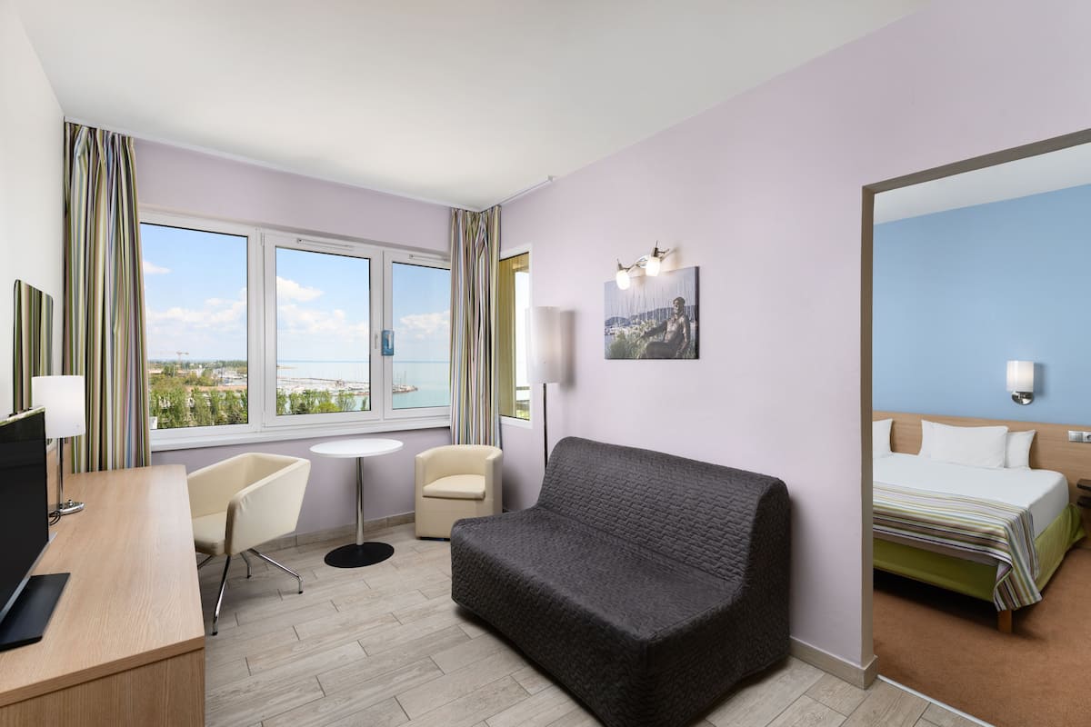 Ein Hotelzimmer mit einem Bett, einem Sofa, einem Schreibtisch mit Computer, einem Stuhl, einem kleinen Tisch und einem Fenster mit Blick auf die Stadt.