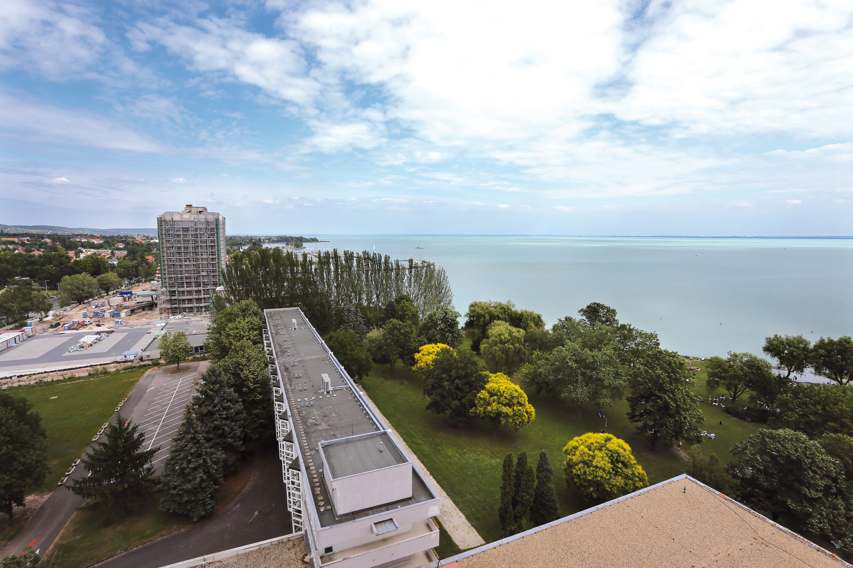 A high-rise building with a rooftop terrace overlooking a body of water and a park with trees.