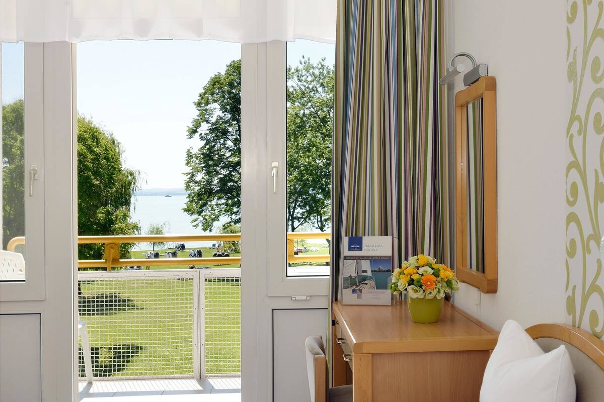 Ein Hotelzimmer mit einem Bett, einem Schreibtisch mit Blumenvase, Blick auf eine Wiese und Wasser durch ein Fenster sowie einer Klimaanlage an der Wand.