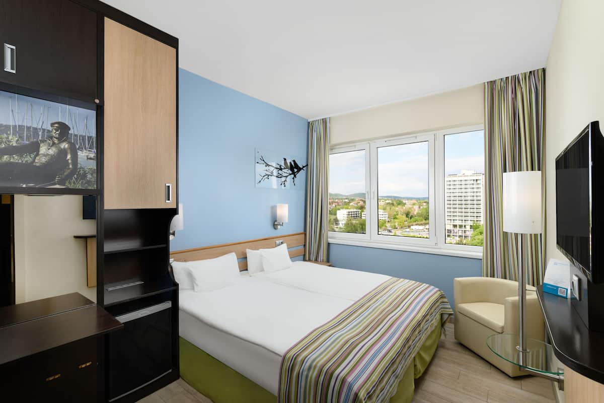Ein Hotelzimmer mit Bett, Schreibtisch, Stuhl, Fernseher und einem Fenster mit Blick auf Gebäude.